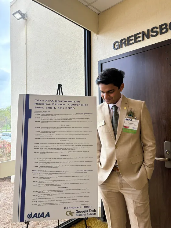 Haitish in a tan suit beside the program board for the 76th AIAA Southeastern Regional Student Conference in Greensboro