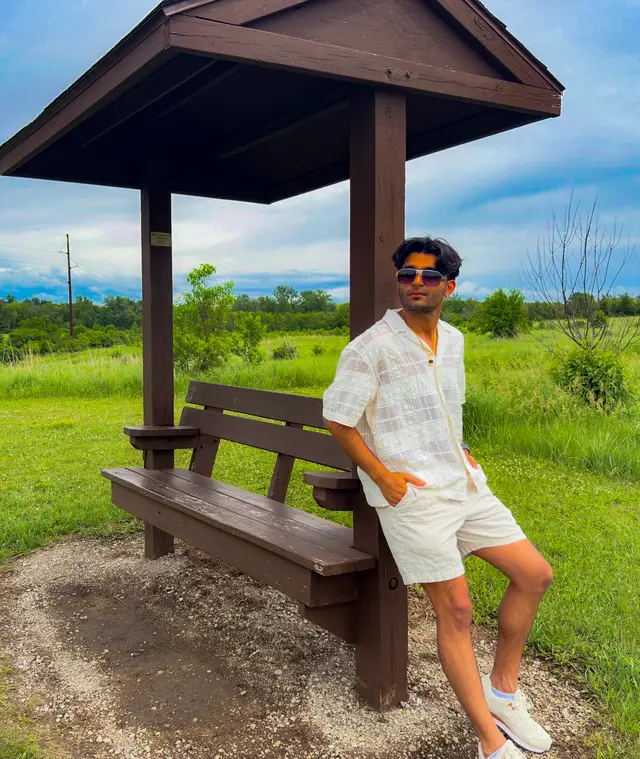 Haitish in sunglasses and white linen leaning on a wooden shelter beside a green field